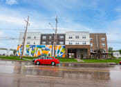 Thumbnail 46 of 58 - A red car is parked in front of a building with a colorful mural on the side.