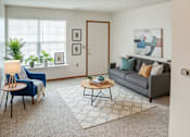 Thumbnail 26 of 29 - A living room with a grey couch, a blue chair, a wooden coffee table, and a rug.