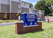 Thumbnail 26 of 31 - A sign for Bluebird Commons apartment complex stands in front of a brick building.