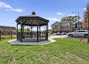 Thumbnail 23 of 31 - A gazebo is situated in a grassy area with a black fence and a car parked in the background.