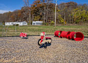 Thumbnail 64 of 76 - A red toy motorcycle is in the foreground of a gravel area with a few red barrels and a few buildings in the background.