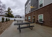 Thumbnail 26 of 37 - A series of black picnic tables are lined up on a concrete patio outside of a brick building.