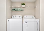 Thumbnail 22 of 22 - Apartment laundry closet with a side-by-side washer and dryer and overhead wire shelving at Canterbury Apartments in Myrtle Beach, SC.