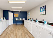 Thumbnail 21 of 22 - Community laundry room with rows of washers and dryers and a folding counter at Canterbury Apartments in Myrtle Beach, SC.