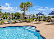 Thumbnail 11 of 22 - Swimming pool steps with a handrail, lounge chairs and shaded tables at Canterbury Apartments in Myrtle Beach, SC.