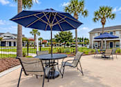Thumbnail 12 of 22 - Poolside tables with navy umbrellas, lounge chairs and palm trees at Canterbury Apartments in Myrtle Beach, SC.