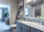 Thumbnail 9 of 28 - Model bathroom with grey sleek cabinets and brushed nickel fittings, granite marbleized looking counter and very large vanity.  Hand towels hanging and artwork behind the toilet. Door leads to bedroom with carpet and window featuring faux wood blinds.