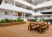 Thumbnail 38 of 63 - A wooden table with benches is in the foreground of a modern building with white balconies.