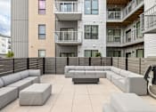 Thumbnail 85 of 88 - A modern outdoor seating area with grey couches and tables is surrounded by apartment buildings.