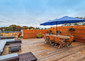 Thumbnail 19 of 36 - A wooden deck with a table and chairs under a blue umbrella.