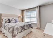 Thumbnail 15 of 20 - Apartments In Aurora, IL - The Apartments At Kirkland Crossing - Bedroom With Bed, Dresser, Carpet Flooring, Nightstand, Lamp, And Window.