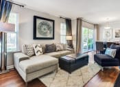 Thumbnail 13 of 20 - Aurora, IL Apartments For Rent - The Apartments At Kirkland Crossing - Living Room With Sectional, Chair, Ottoman, Wood-Style Flooring, Modern Decor, Area Rug, Sliding Glass Door To Back, And Floor Lamps.