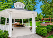 Thumbnail 3 of 18 - A white gazebo with a black roof and a white railing is surrounded by green bushes.