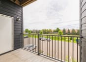 Thumbnail 29 of 56 - A balcony with a metal railing, overlooking a parking lot and green trees in the distance. The view is framed by the building's exterior wall and a door.