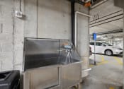 Thumbnail 31 of 56 - A stainless steel utility sink located in a parking garage, with a hose and a trash can nearby. A white vehicle is visible in the background.