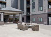 Thumbnail 39 of 56 - An outdoor seating area with a wicker sofa set, including chairs and a coffee table, located under a covered pavilion. The space is surrounded by modern apartment buildings.