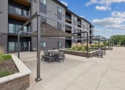 Thumbnail 41 of 56 - An outdoor seating area with shaded tables under canopies, surrounded by landscaped planters and modern apartment buildings. The area features a paved pathway and clear blue skies in the background.