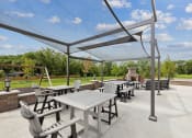 Thumbnail 37 of 56 - An outdoor patio area with shaded seating, including tables and chairs, under a canopy. The space features a view of green trees and a swing set in the distance.