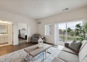 Thumbnail 15 of 56 - A bright living room with a sofa, coffee table, and decorative items. The space features large windows and a sliding glass door leading to an outdoor area, with a glimpse of the adjacent room visible in the background.