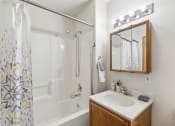 Thumbnail 15 of 31 - Modern apartment bathroom with a white bathtub and shower combination, a patterned shower curtain, and a vanity with a wooden cabinet and mirrored medicine cabinet. The space features bright vanity lighting, a grab bar for accessibility, and neatly arranged toiletries
