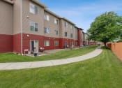 Thumbnail 17 of 31 - Three-story apartment building with beige and red siding, featuring large windows and patio doors on the ground level. A curved walking path winds through the well-manicured lawn, lined with mature trees and a wooden privacy fence
