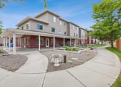 Thumbnail 16 of 31 - Three-story apartment building with beige and red siding, featuring a covered patio area with outdoor seating. The landscaped courtyard includes a walking path, decorative garden features, and raised garden beds, surrounded by lush greenery and mature trees
