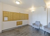 Thumbnail 31 of 31 - Apartment mailroom featuring a wall of gold-colored mailboxes, two gray upholstered chairs, and warm lighting. The space has wood-style flooring and a neutral color palette, creating a clean and inviting environment