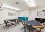 Thumbnail 25 of 31 - Community game and activity room featuring a poker table with cup holders, a long table with board games, and teal upholstered chairs along the wall. The space has wood-style flooring, bright overhead lighting, and modern decor for a welcoming atmosphere