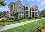 Thumbnail 2 of 17 - A large apartment complex with a well-maintained lawn and flower beds in front.