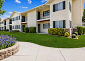 Thumbnail 2 of 22 - A building with a white facade and balconies is surrounded by a well-manicured lawn and a stone planter with purple flowers.