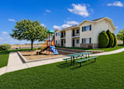 Thumbnail 5 of 22 - A playground with a slide and a bench is in front of a two-story building.