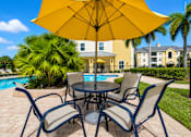 Thumbnail 6 of 24 - A table with chairs and an umbrella is in the foreground of a pool area.