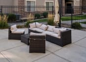 Thumbnail 17 of 31 - Outdoor lounge area in an apartment community featuring a wicker sectional sofa with beige cushions, a matching armchair, and a glass-top coffee table. The space is set on a paved patio with decorative grasses, a closed patio umbrella, and a black metal fence enclosing the area