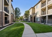 Thumbnail 11 of 31 - Modern apartment community with three-story buildings featuring brick and stucco exteriors. A winding concrete walkway leads through well-manicured green lawns and mature trees, creating a peaceful residential setting