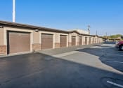 Thumbnail 30 of 31 - Row of private garage units in an apartment community, featuring brown roll-up doors, beige stucco walls, and brick accents. The area is paved with asphalt and offers secure, enclosed parking for residents