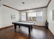 Thumbnail 28 of 31 - Modern apartment clubhouse game room featuring a black pool table, wooden flooring, and large windows with sheer curtains. The space includes a sleek overhead light fixture, abstract wall art, and a cue rack for pool accessories