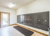 Thumbnail 29 of 31 - Apartment community mailroom featuring a wall of black metal mailboxes, a tiled floor with a dark entry mat, and a glass door leading outside. The well-lit space provides residents with secure and organized mail storage