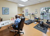 Thumbnail 9 of 22 - A woman in a blue shirt is sitting at a table in a room with exercise equipment.