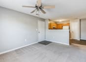 Thumbnail 1 of 26 - Empty living room with light gray walls and carpeted flooring, featuring a ceiling fan with wooden blades. The room opens into a kitchen with wooden cabinets, white appliances, and a breakfast bar. A white door leads to another area of the home, and there is a mix of carpet and hardwood flooring