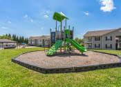 Thumbnail 24 of 26 - Outdoor playground with green slides, climbing features, and a shaded platform, surrounded by a mulch-filled play area with a black border. The playground is situated on a grassy lawn near multi-story apartment buildings, with a clear blue sky overhead. A swimming pool area with lounge chairs is visible in the background