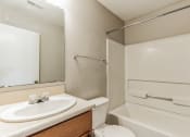 Thumbnail 18 of 26 - Bathroom with a white bathtub and shower combination, featuring a built-in grab bar and a shower rod above. A vanity with a white sink, wooden cabinet, and beige countertop is next to a toilet. A large mirror is mounted above the sink, reflecting a white door. The walls are painted in a neutral tone, and a towel rack is installed on the side wall