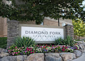 Thumbnail 1 of 9 - A sign for Diamond Fork Apartments is surrounded by flowers and greenery. at Diamond Fork Apartments, Apartments, Spanish Fork, UT