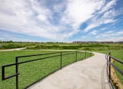 Thumbnail 18 of 25 - A concrete walkway with a metal railing leads through a grassy field.