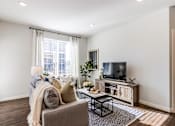 Thumbnail 3 of 25 - A living room with a grey couch, a coffee table, a television, and a window with white curtains.
