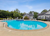 Thumbnail 12 of 17 - Luxury apartment community outdoor pool with crystal-clear water, surrounded by a spacious sundeck with lounge chairs. A brick and white clubhouse with large windows and landscaping is in the background, along with lush green trees under a bright blue sky