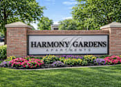 Thumbnail 1 of 28 - A sign for Harmony Gardens Apartments is framed by flowers and topped by two pillars.
