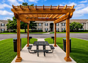 Thumbnail 6 of 12 - A wooden pergola with a picnic table is situated in a grassy area in front of apartment buildings.
