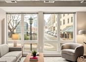 Thumbnail 3 of 43 - A living room with a grey couch, a grey armchair, a wooden table with a red book and a plant on it, and a view of a street through the windows.
