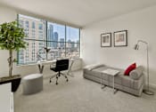 Thumbnail 3 of 40 - A room with a grey couch, a desk with a chair, a potted plant, and two framed pictures on the wall.