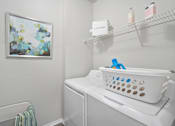 Thumbnail 13 of 42 - A laundry room with a white washer and dryer, a basket on top of the dryer, and a painting on the wall at The Grand Reserve at Tampa Palms Apartments, Tampa, FL, 33647